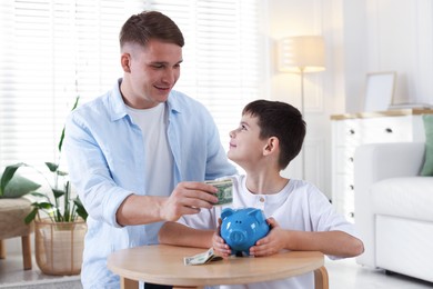 Photo of Father and son putting pocket money into piggybank at table indoors