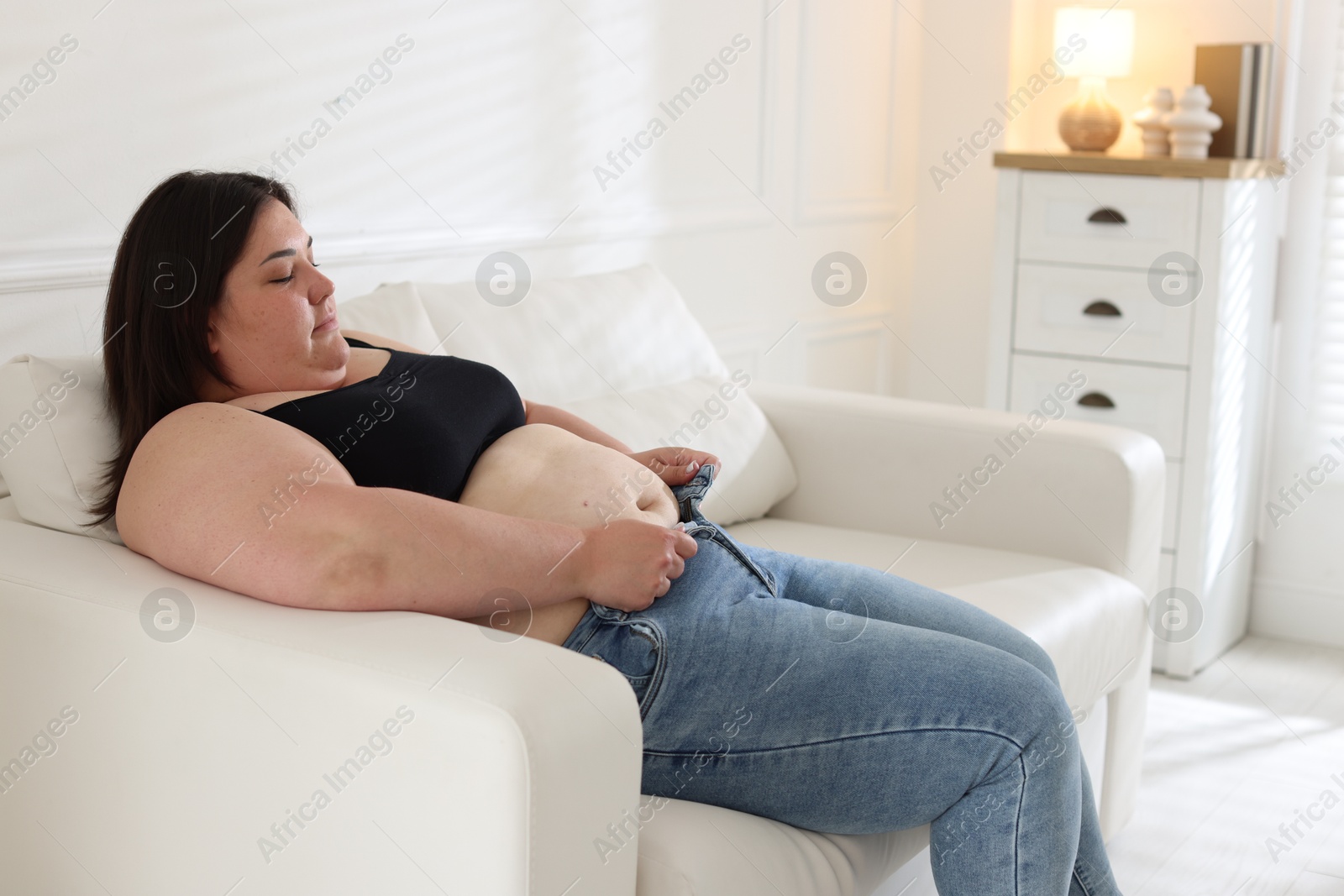 Overweight woman trying to put on tight jeans indoors Photo of Overweight woman trying to put on tight jeans indoors