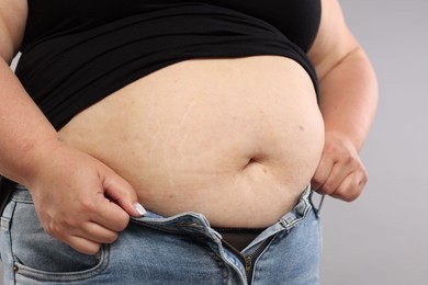 Photo of Overweight woman trying to put on tight jeans against grey background, closeup