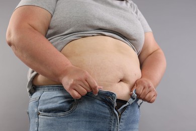 Photo of Overweight woman trying to put on tight jeans against grey background, closeup
