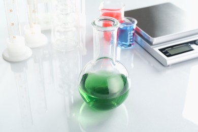 Photo of Flask with green liquid, other laboratory glassware and scale on white mirror table, closeup