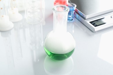 Photo of Flask with green liquid, other laboratory glassware and scale on white mirror table, closeup