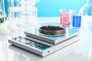 Photo of Scale with Petri dish of sample and other laboratory glassware on white mirror table against light blue background, closeup