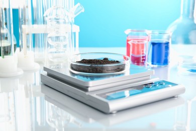 Photo of Scale with Petri dish of sample and other laboratory glassware on white mirror table against light blue background, closeup