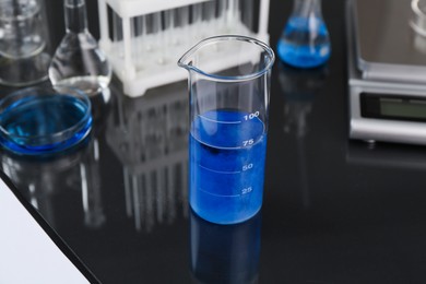 Photo of Beaker with blue liquid, other laboratory glassware and scale on black mirror table, closeup