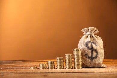 Photo of Bag with dollar sign and stacked coins on wooden table against brown background, space for text