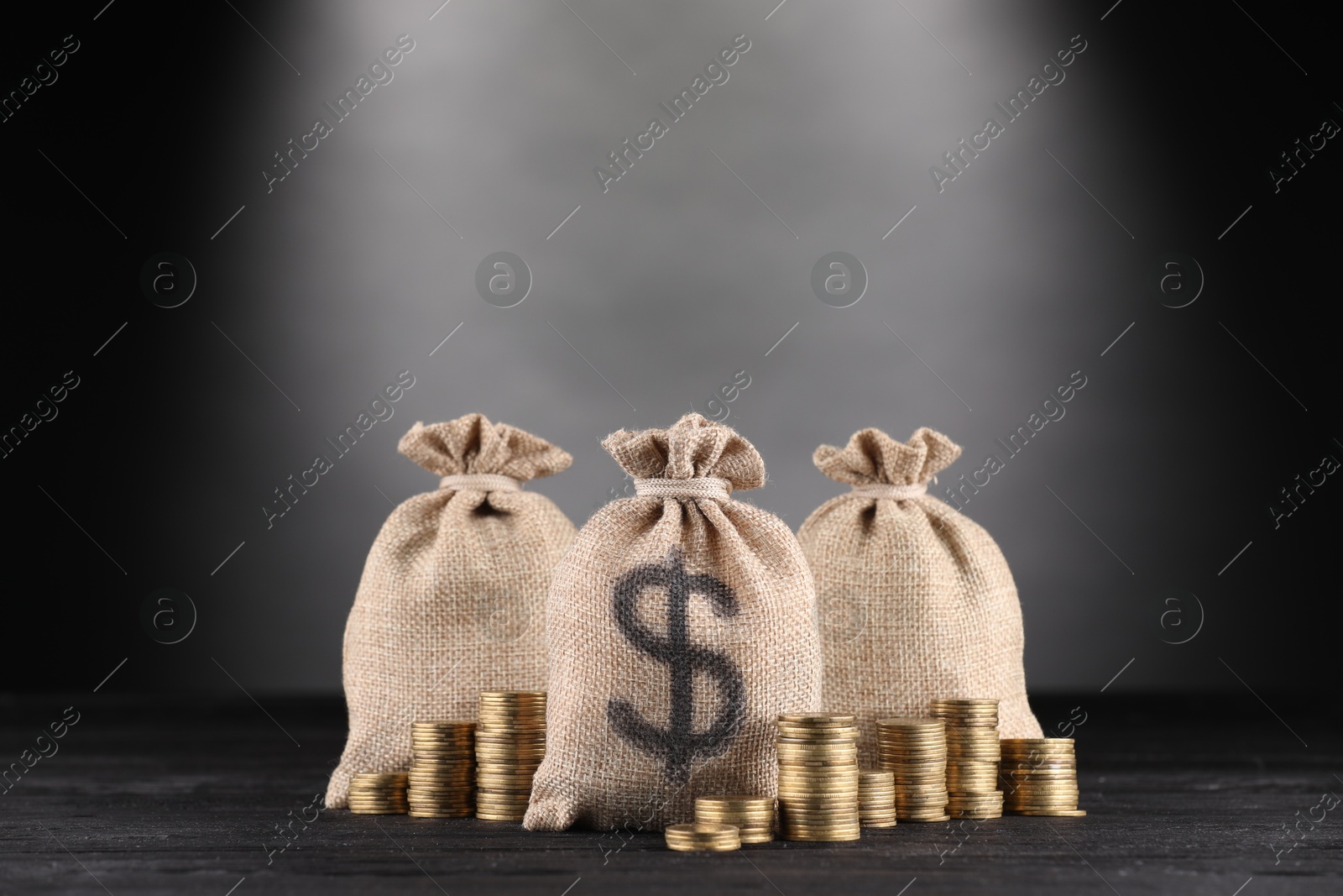 Photo of Bags with dollar sign and stacked coins on black wooden table against gray gradient background