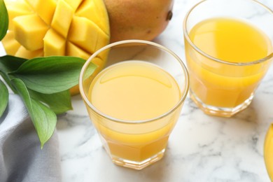 Photo of Tasty mango juice in glasses, fresh fruits and green leaves on white marble table, closeup