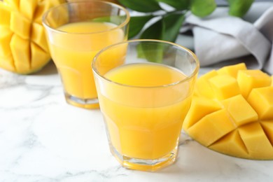 Photo of Tasty mango juice in glasses, fresh fruits and green leaves on white marble table, closeup