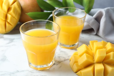Photo of Tasty mango juice in glasses, fresh fruits and green leaves on white marble table, closeup