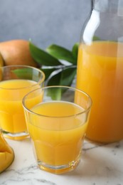 Photo of Tasty mango juice in glasses, fresh fruit and green leaves on white marble table, closeup