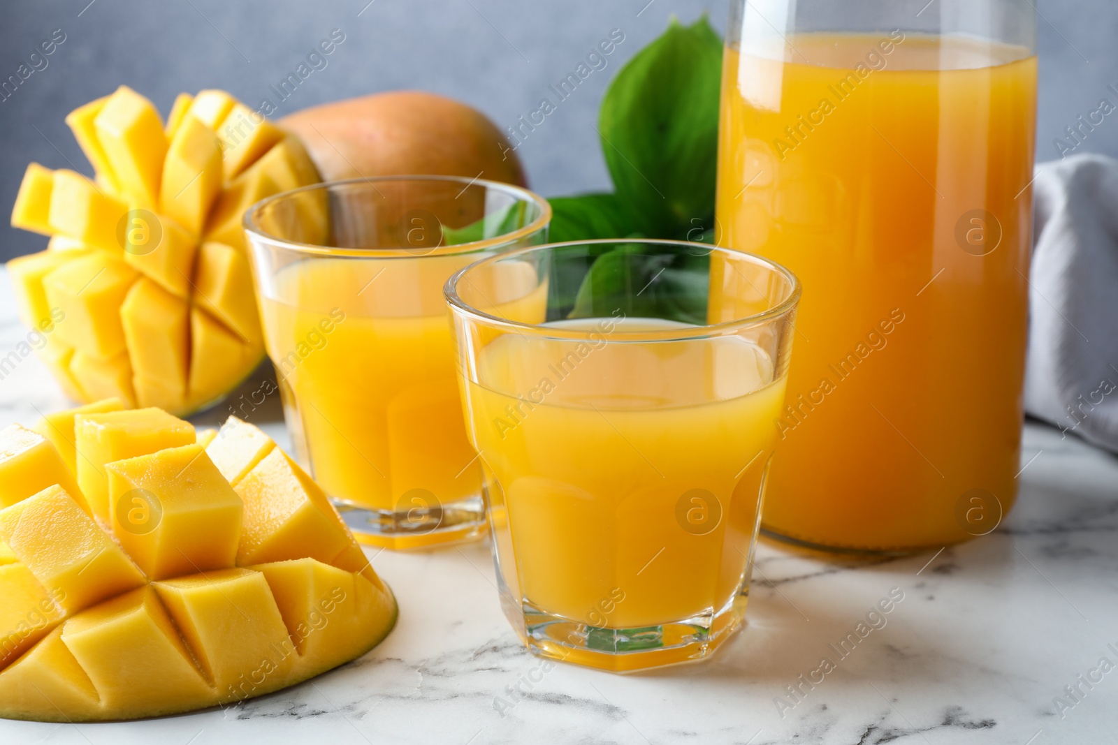Tasty mango juice in glasses, fresh fruits and green leaves on white marble table, closeup Photo of Tasty mango juice in glasses, fresh fruits and green leaves on white marble table, closeup