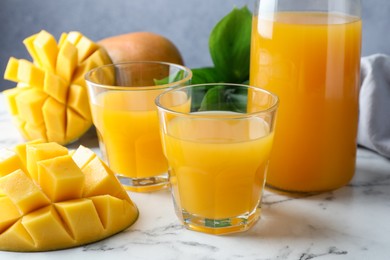 Photo of Tasty mango juice in glasses, fresh fruits and green leaves on white marble table, closeup