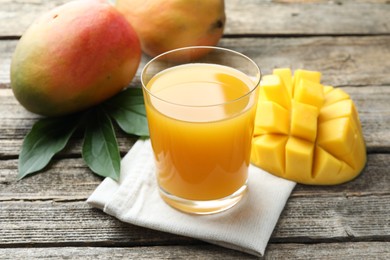 Photo of Tasty mango juice in glass, fresh fruits and green leaves on wooden table, closeup