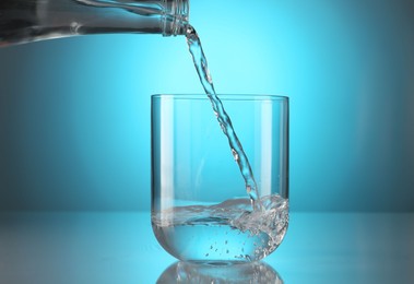 Photo of Pouring water from bottle into glass on mirror table against light blue gradient background. Backlit