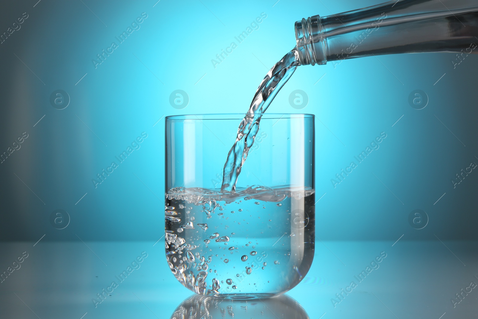Pouring water from bottle into glass on mirror table against light blue gradient background. Backlit Photo of Pouring water from bottle into glass on mirror table against light blue gradient background. Backlit