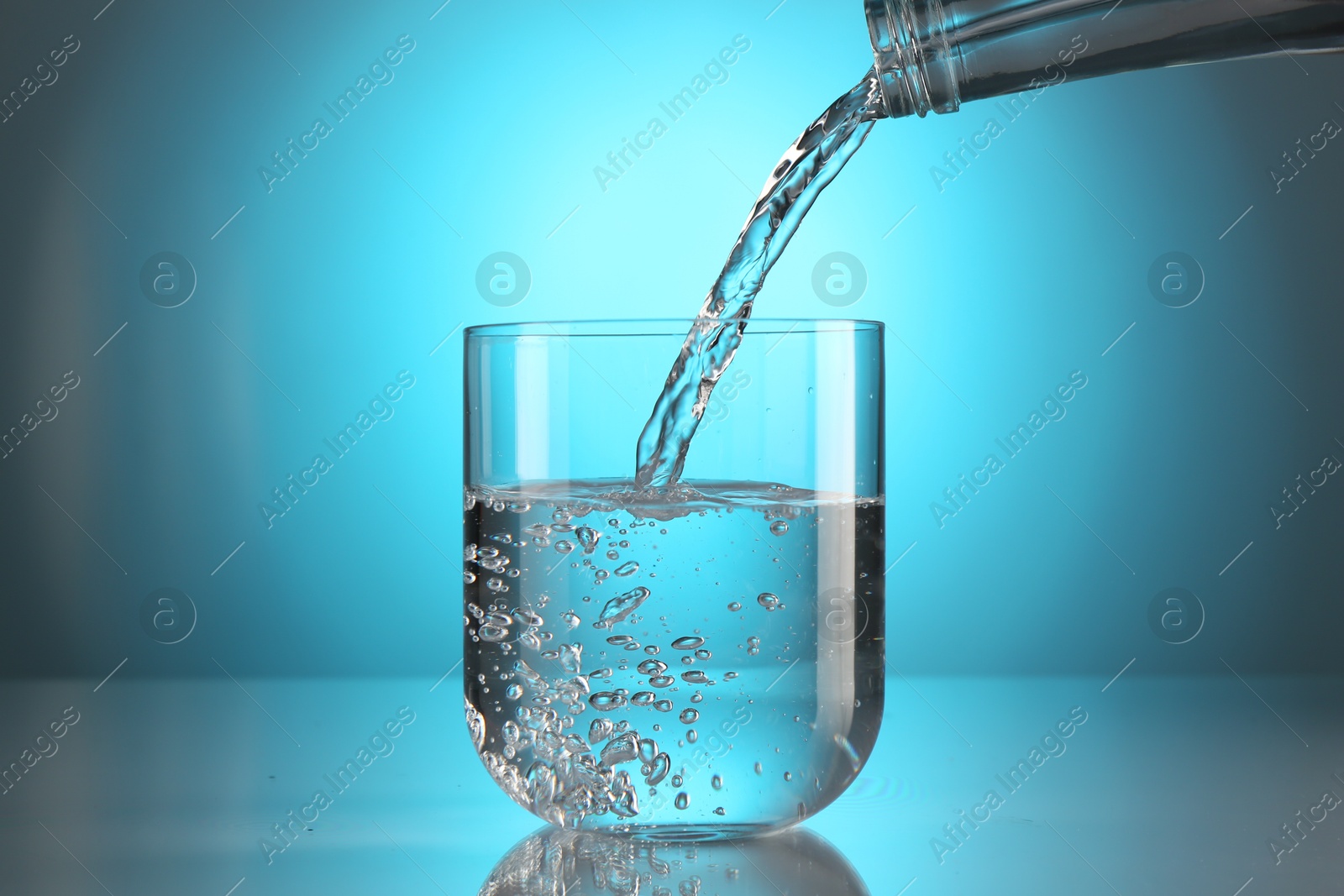 Pouring water from bottle into glass on mirror table against light blue gradient background. Backlit Photo of Pouring water from bottle into glass on mirror table against light blue gradient background. Backlit