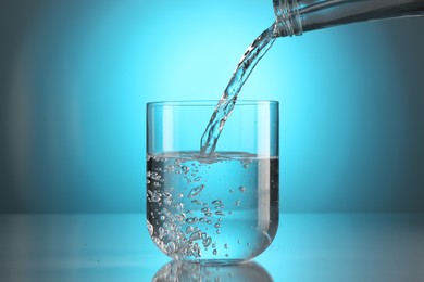 Photo of Pouring water from bottle into glass on mirror table against light blue gradient background. Backlit