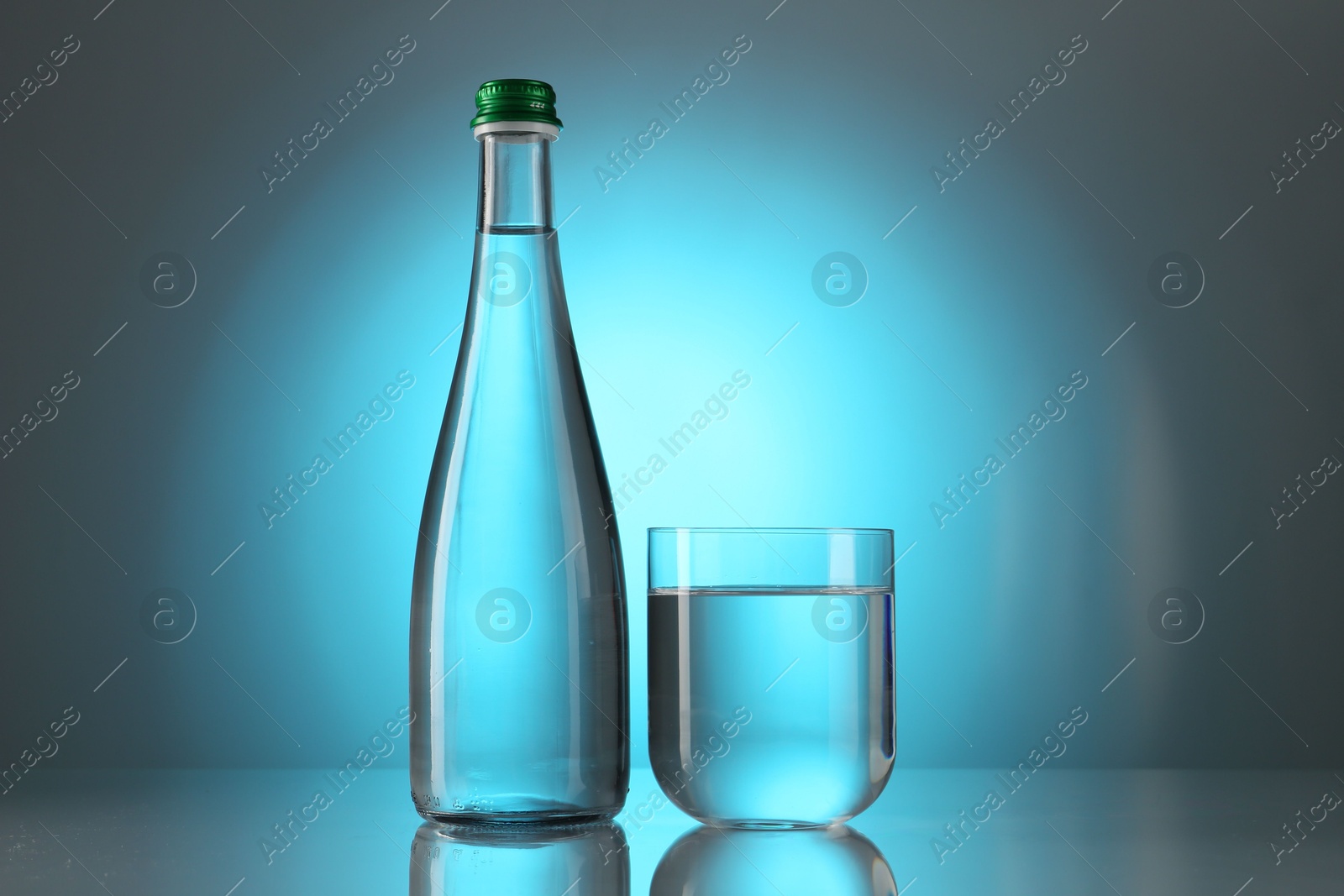 Photo of Fresh water in glass and bottle on mirror table against light blue gradient background. Backlit