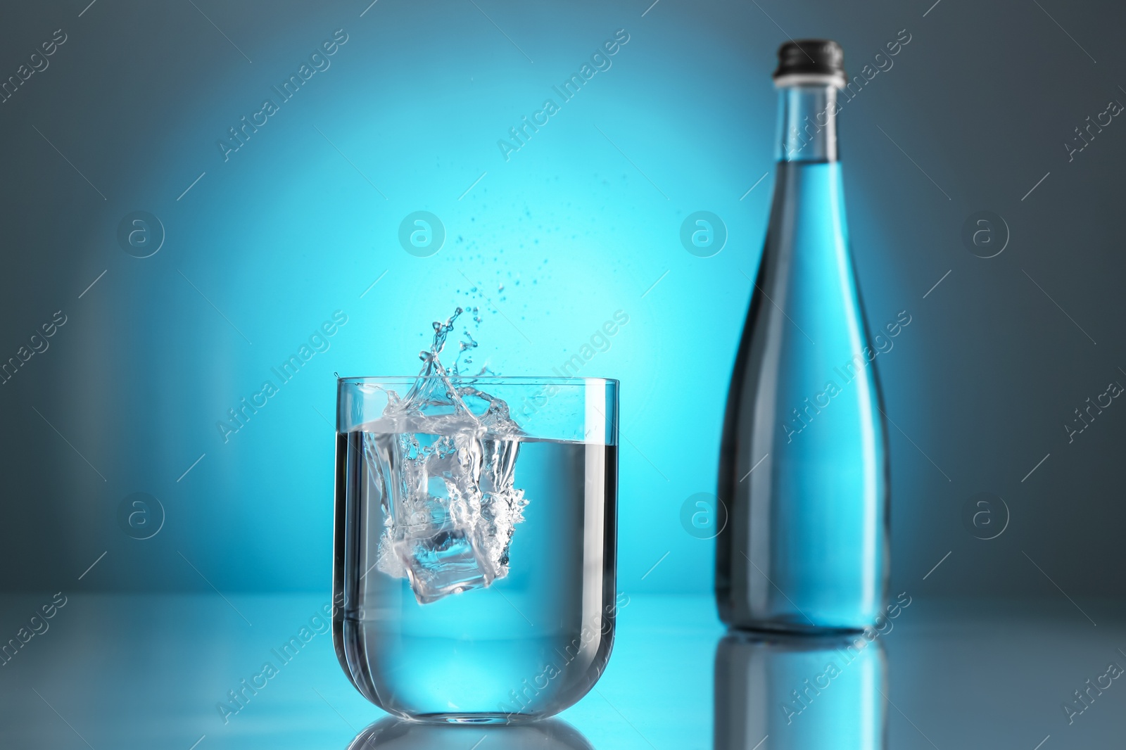 Fresh water splashing out of glass on mirror table against light blue gradient background. Backlit Photo of Fresh water splashing out of glass on mirror table against light blue gradient background. Backlit