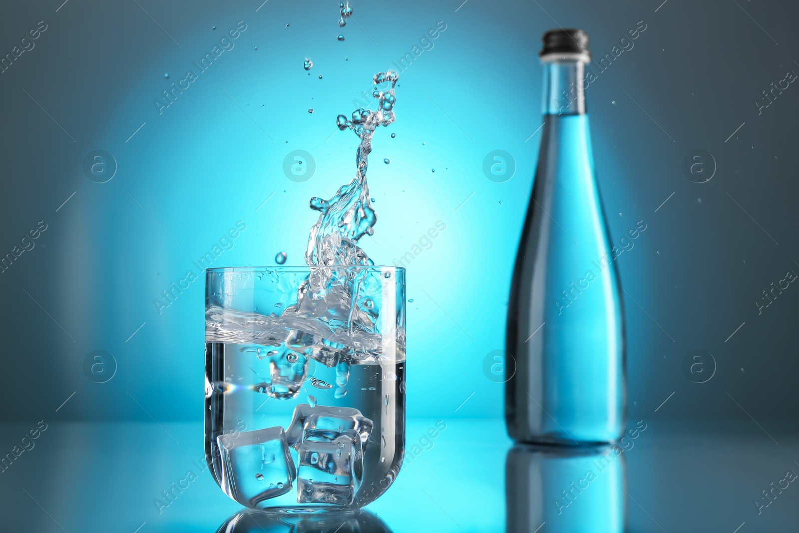 Fresh water splashing out of glass on mirror table against light blue gradient background. Backlit Photo of Fresh water splashing out of glass on mirror table against light blue gradient background. Backlit