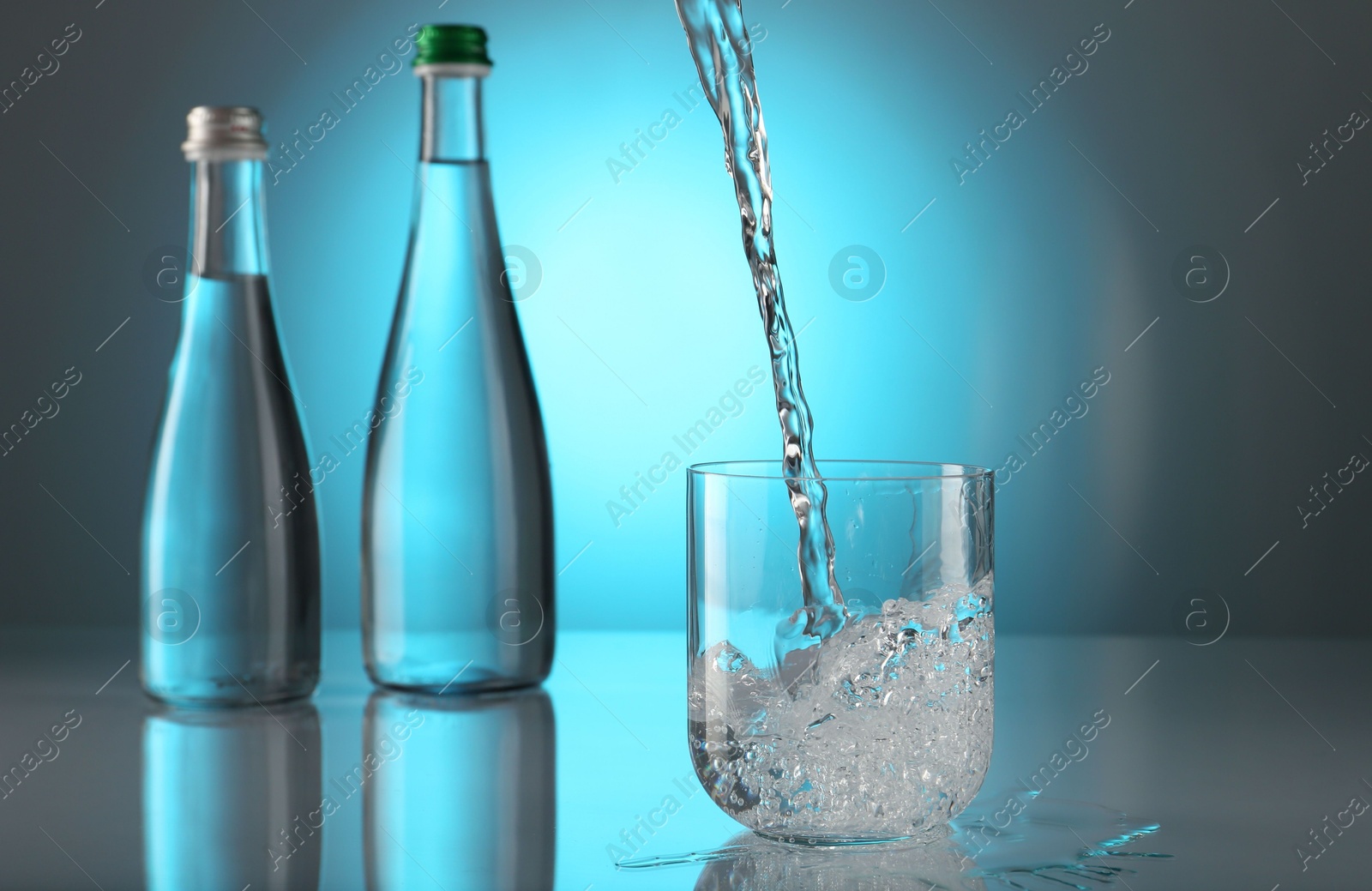 Photo of Pouring water into glass on mirror table against light blue gradient background. Backlit