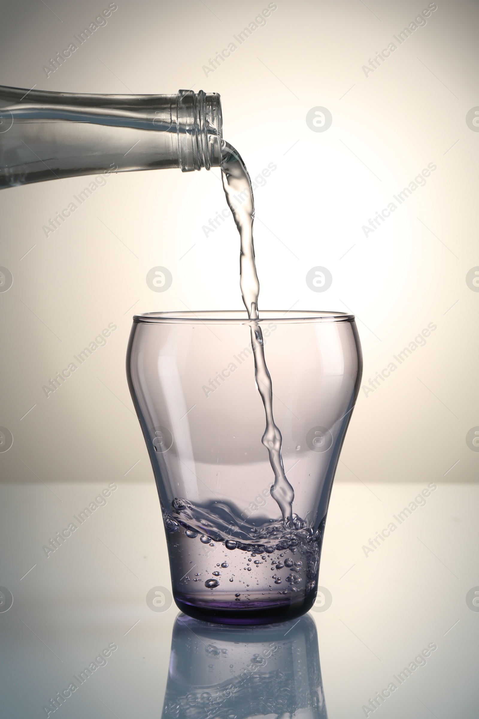 Pouring water from bottle into glass on mirror table against gray gradient background Photo of Pouring water from bottle into glass on mirror table against gray gradient background