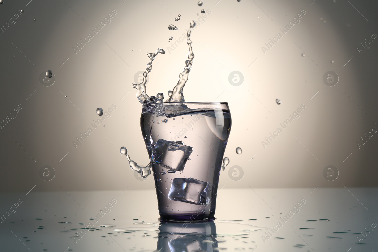 Fresh water splashing out of glass on mirror table against gray gradient background Photo of Fresh water splashing out of glass on mirror table against gray gradient background
