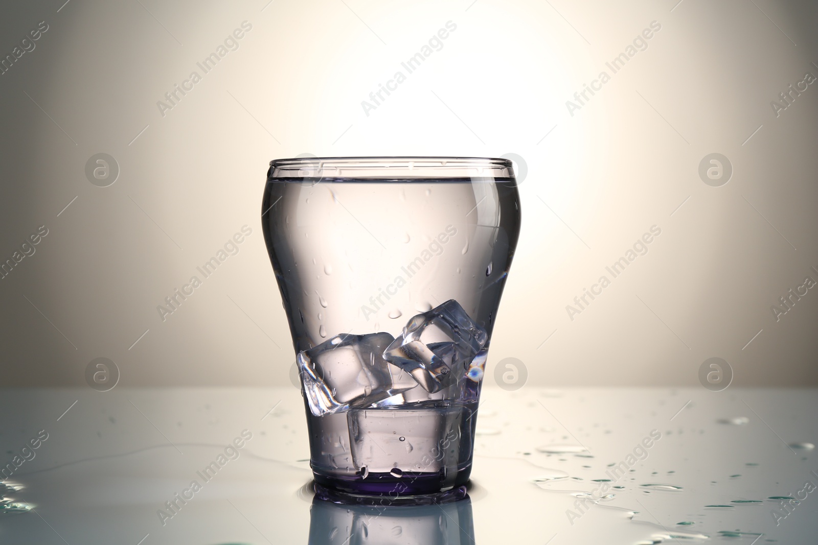 Photo of Fresh water with ice cubes in glass on mirror table against gray gradient background