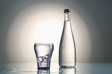 Photo of Fresh water with ice cubes in glass and bottle on mirror table against gray gradient background
