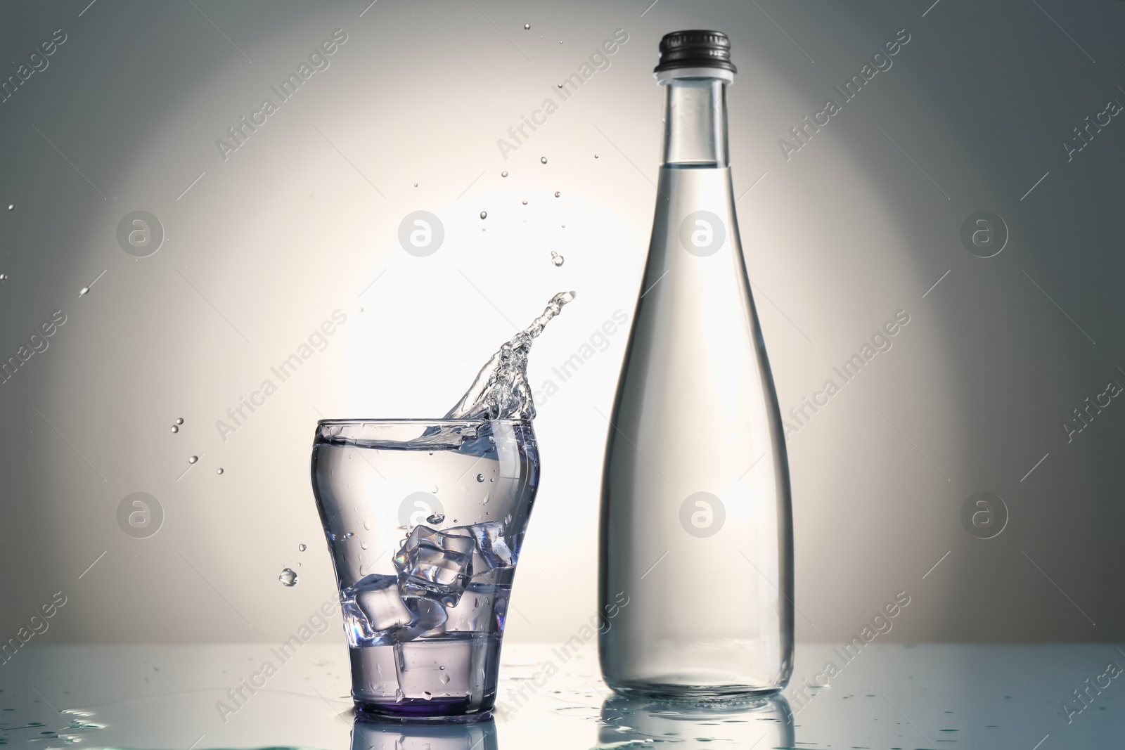 Fresh water splashing out of glass on mirror table against gray gradient background Photo of Fresh water splashing out of glass on mirror table against gray gradient background