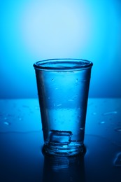Photo of Fresh water with ice cube in glass on mirror table against blue gradient background. Backlit