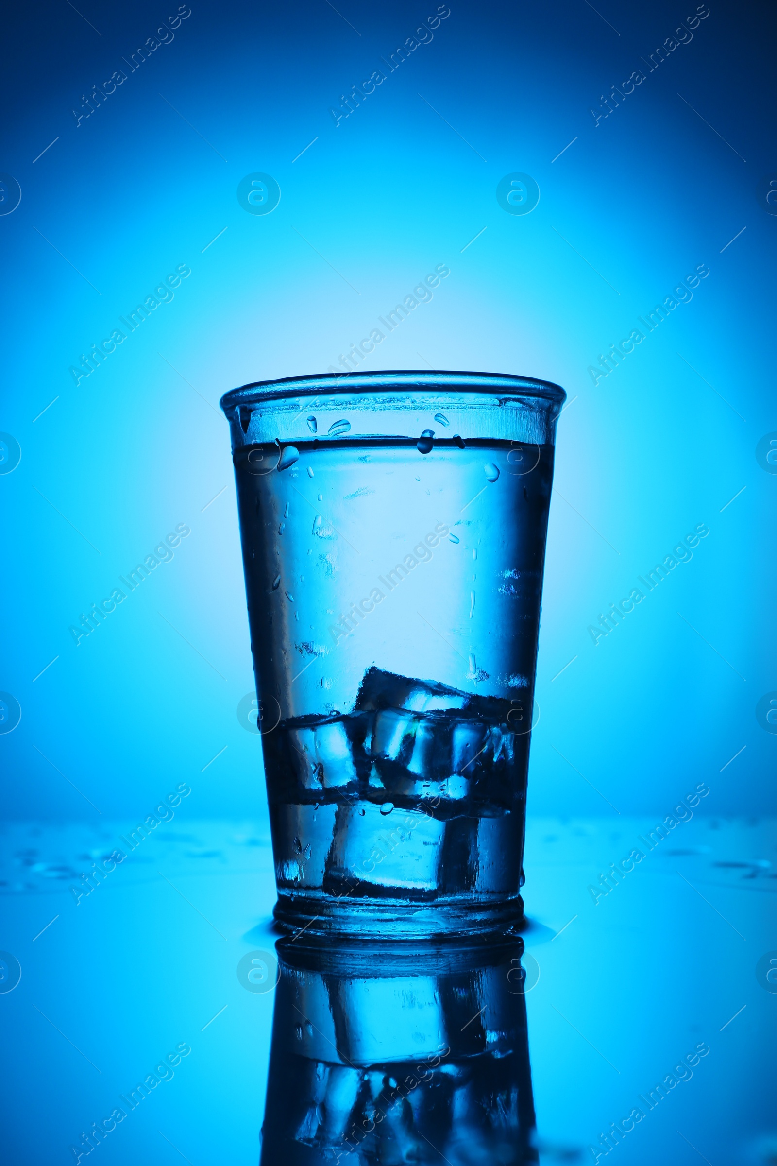 Fresh water with ice cubes in glass on mirror table against blue gradient background. Backlit Photo of Fresh water with ice cubes in glass on mirror table against blue gradient background. Backlit