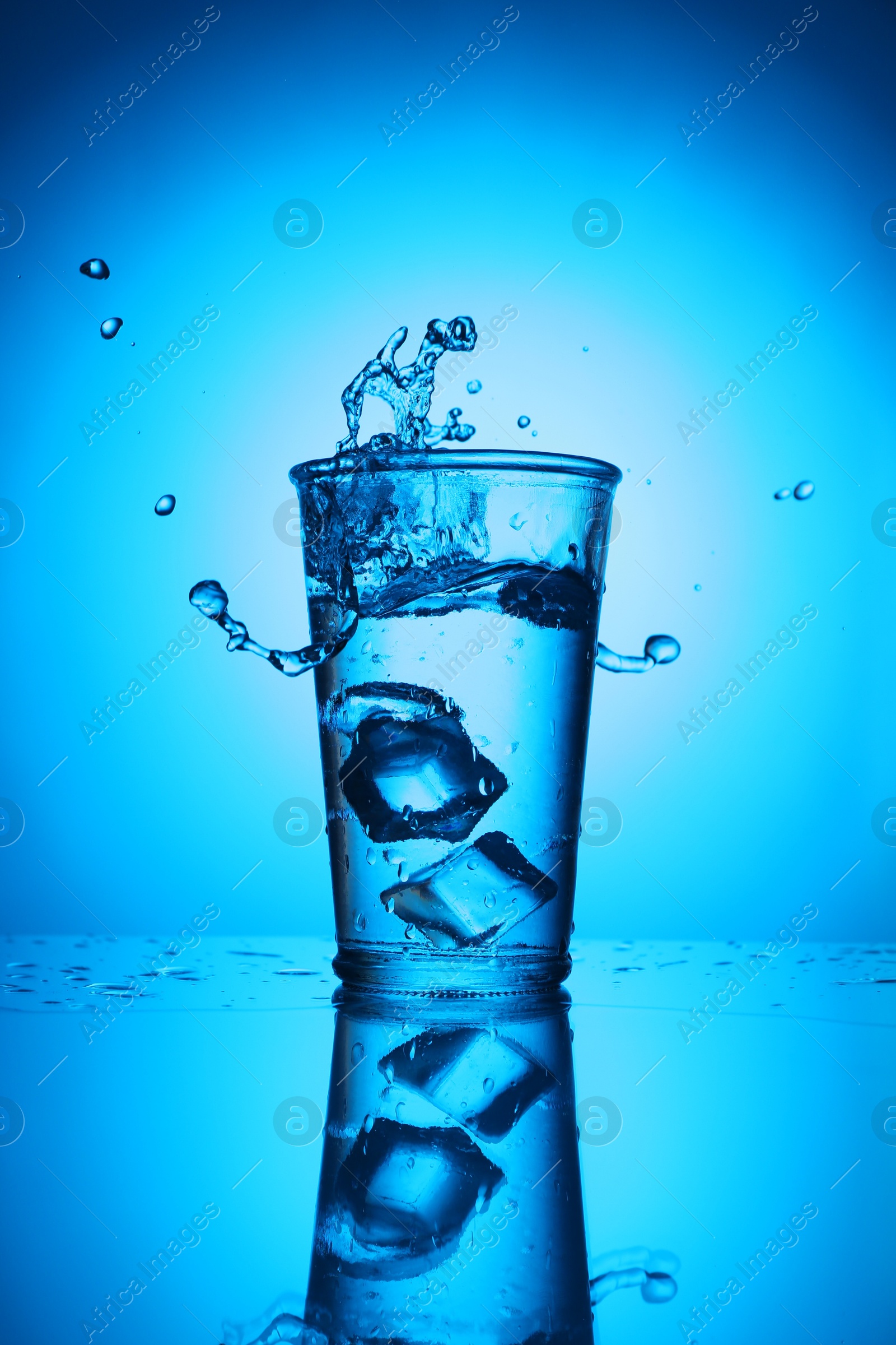 Fresh water with ice cubes in glass on mirror table against blue gradient background. Backlit Photo of Fresh water with ice cubes in glass on mirror table against blue gradient background. Backlit