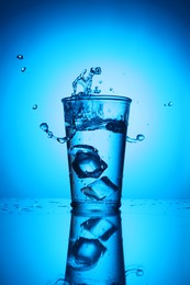 Photo of Fresh water with ice cubes in glass on mirror table against blue gradient background. Backlit