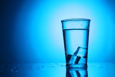 Photo of Fresh water with ice cubes in glass on mirror table against blue gradient background, space for text. Backlit