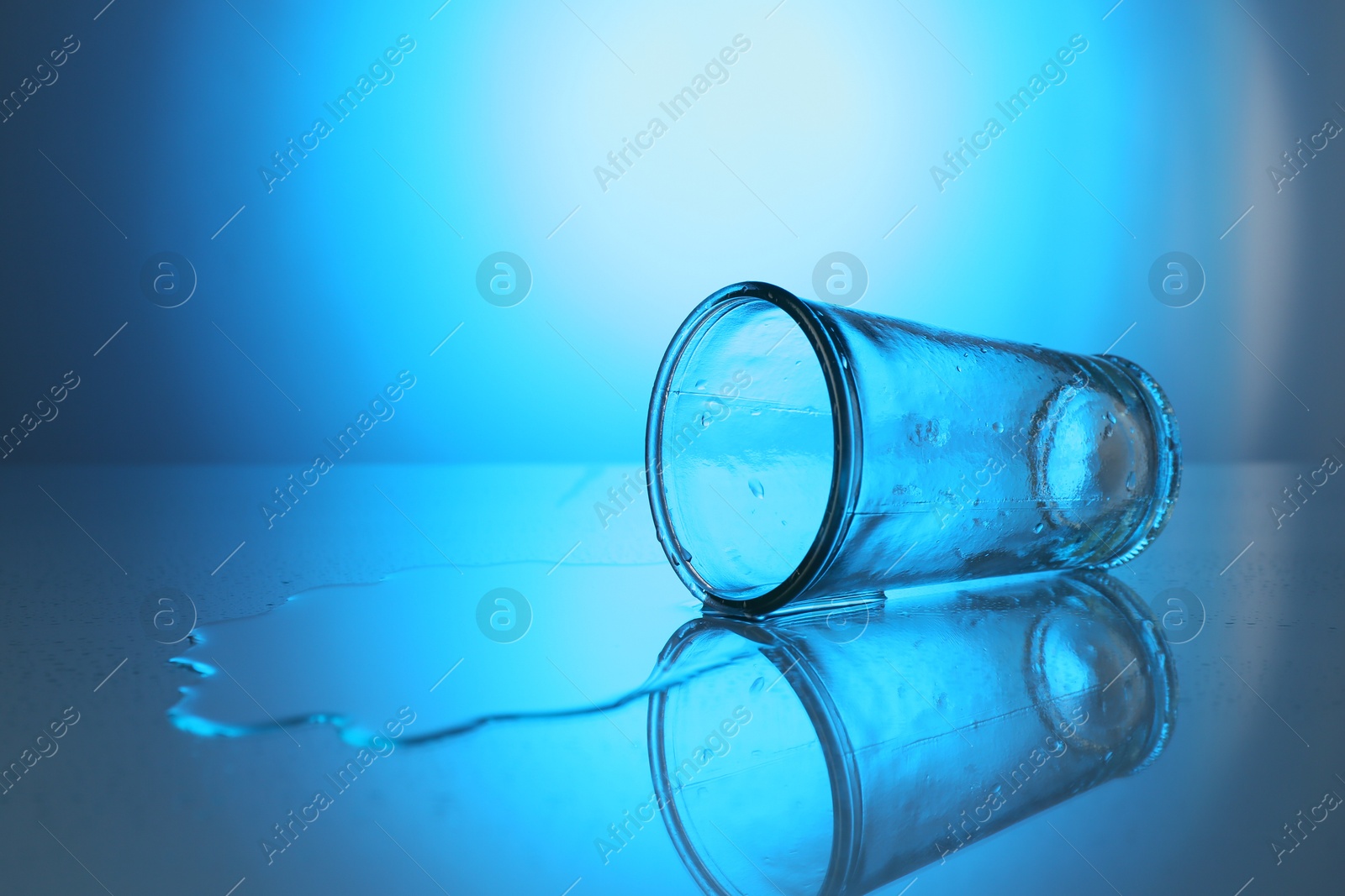 Overturned glass and spilled water on mirror table against blue gradient background, space for text. Backlit Photo of Overturned glass and spilled water on mirror table against blue gradient background, space for text. Backlit