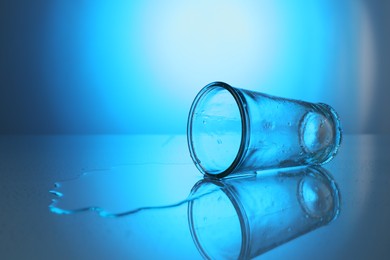 Photo of Overturned glass and spilled water on mirror table against blue gradient background, space for text. Backlit