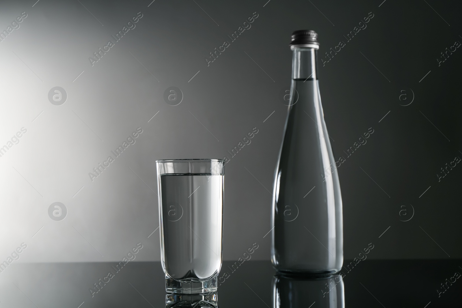 Fresh water in glass and bottle on mirror table against gray background Photo of Fresh water in glass and bottle on mirror table against gray background