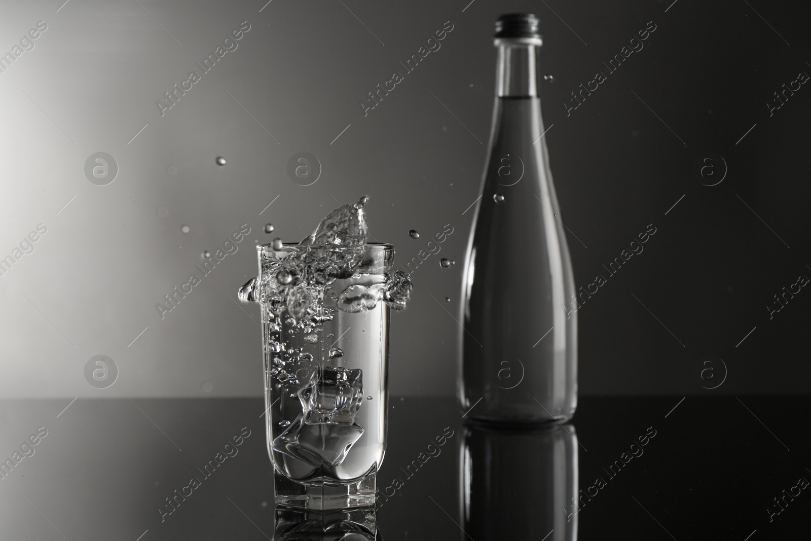Fresh water splashing out of glass on mirror table against gray background Photo of Fresh water splashing out of glass on mirror table against gray background