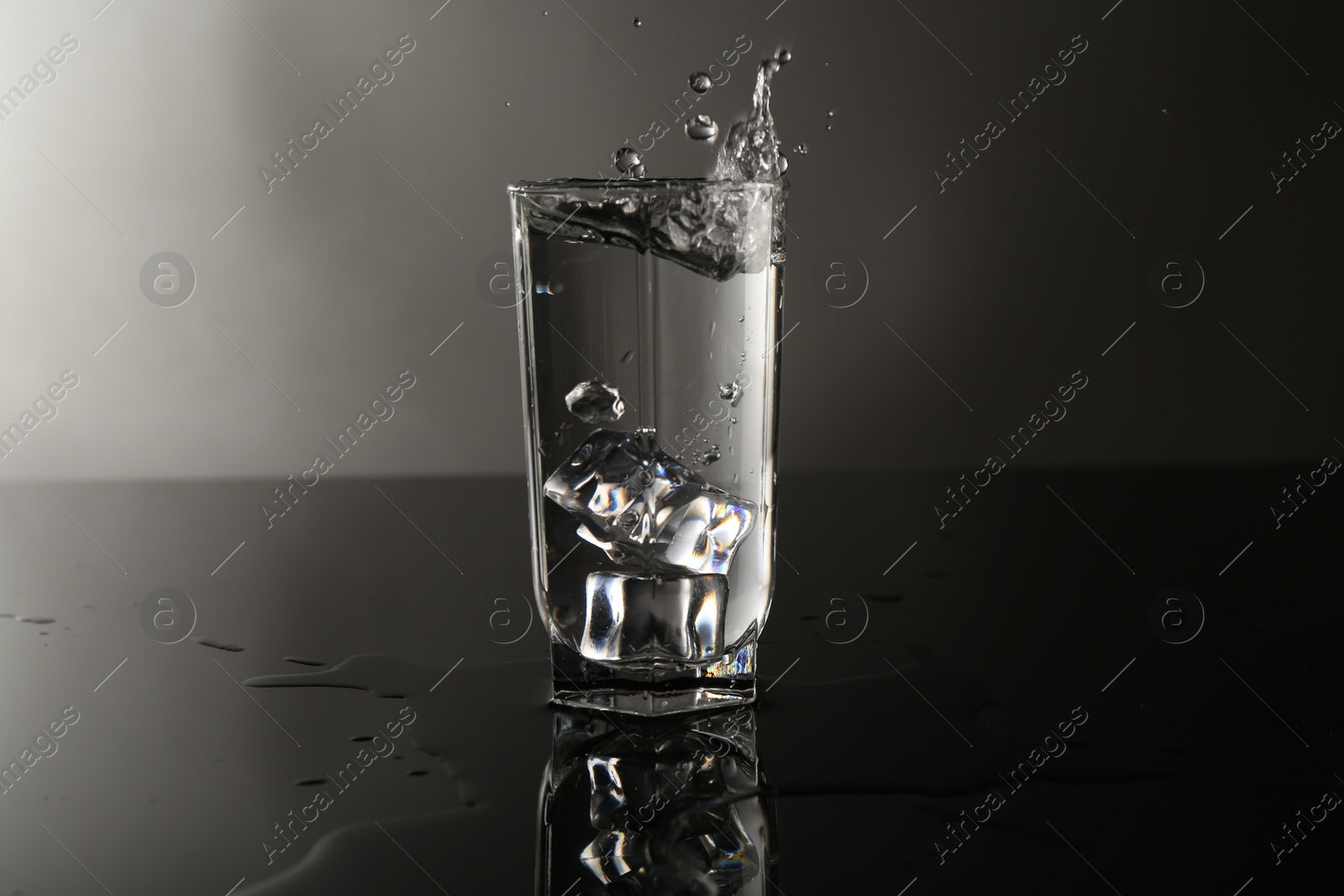Photo of Fresh water splashing out of glass on mirror table against gray background