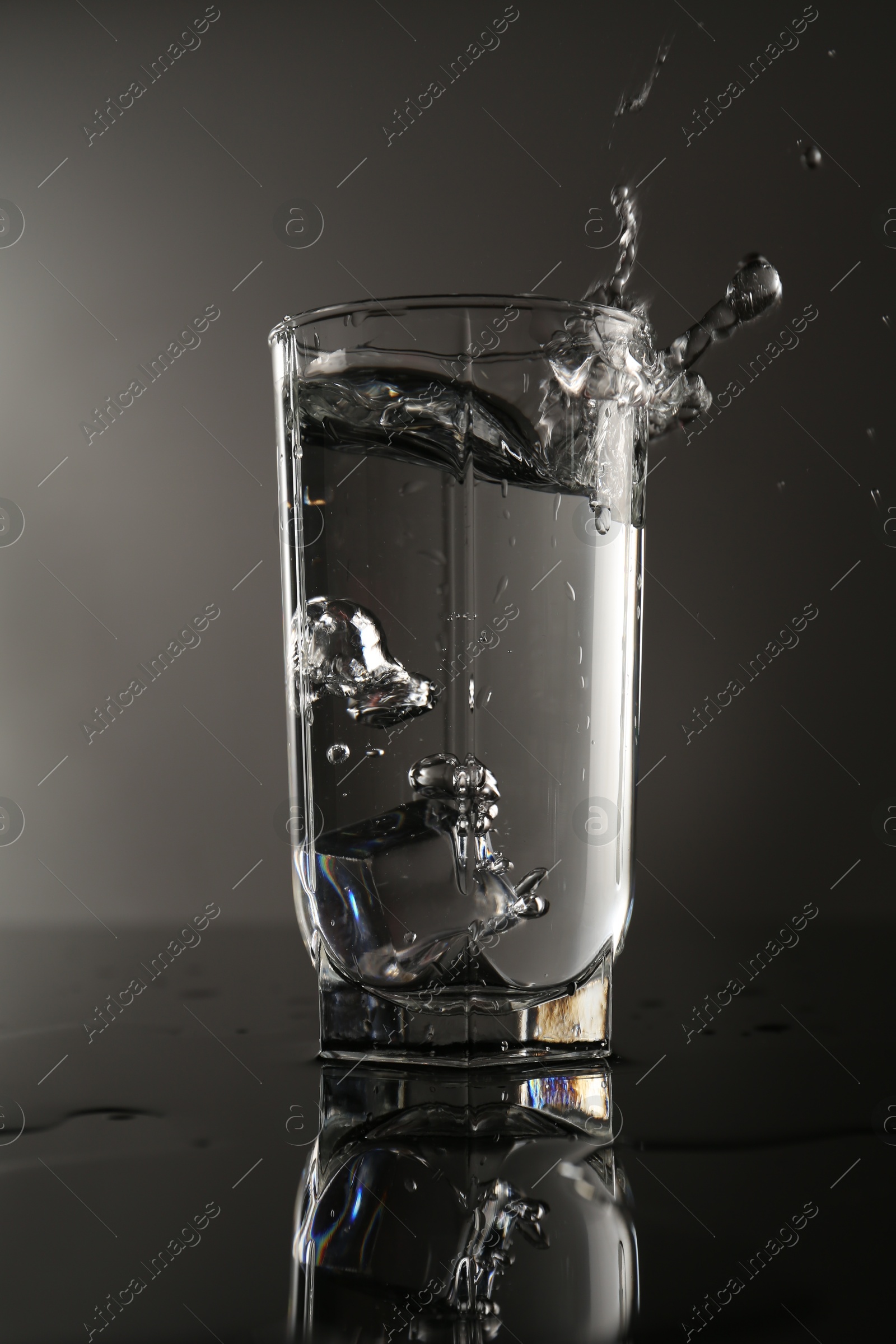Fresh water splashing out of glass on mirror table against gray background Photo of Fresh water splashing out of glass on mirror table against gray background