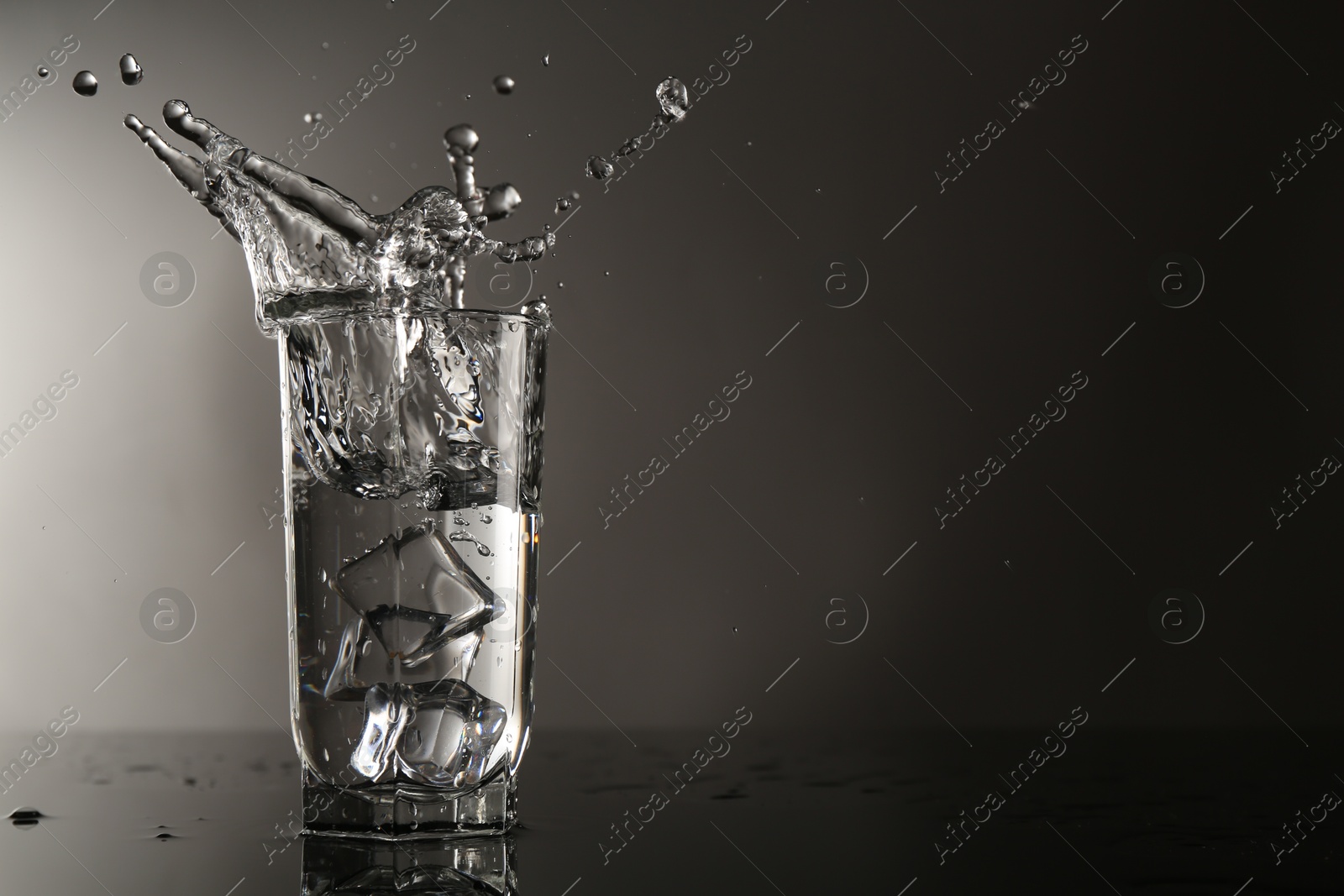Fresh water splashing out of glass on mirror table against gray background, space for text Photo of Fresh water splashing out of glass on mirror table against gray background, space for text