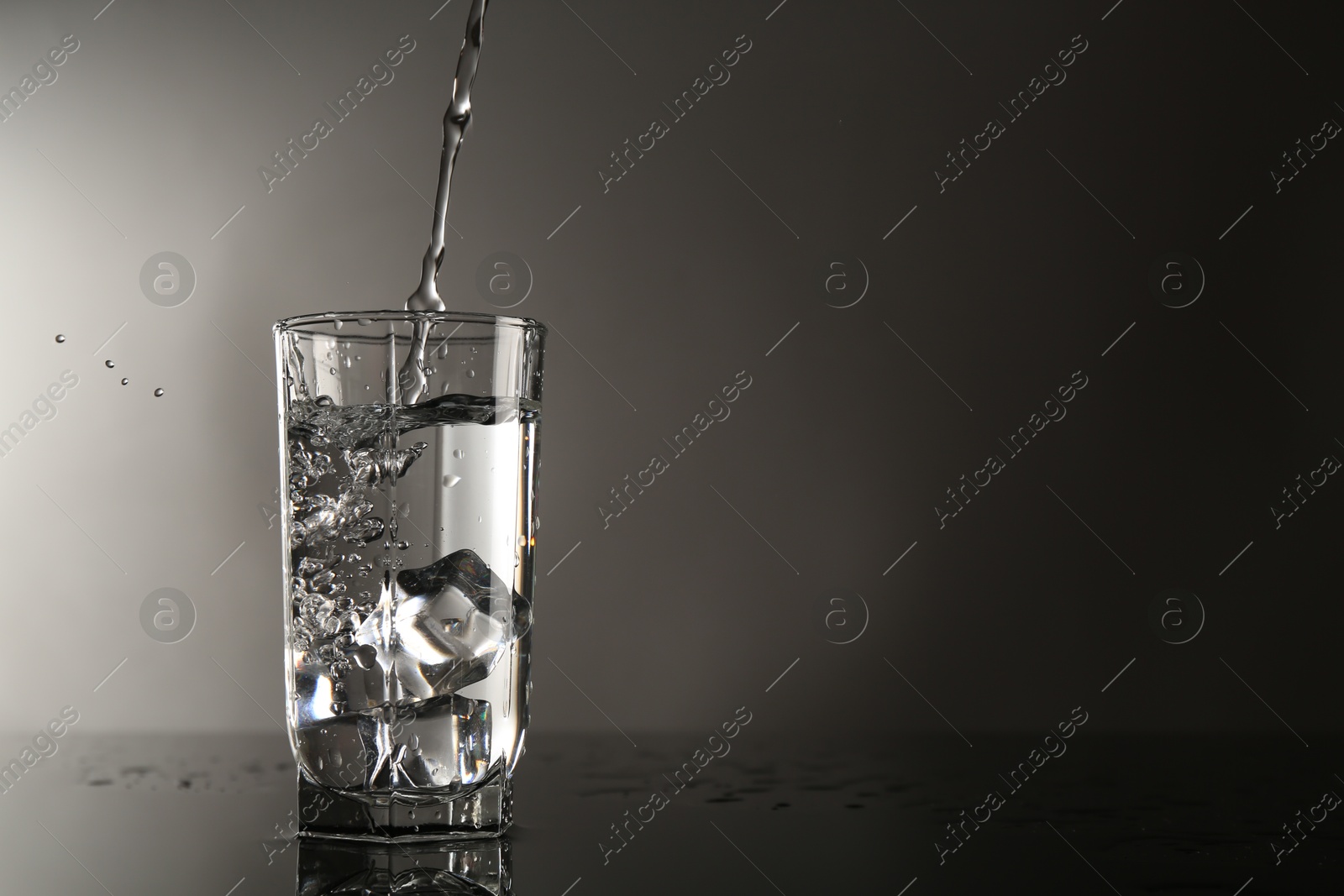 Pouring water into glass on mirror table against gray background, space for text Photo of Pouring water into glass on mirror table against gray background, space for text