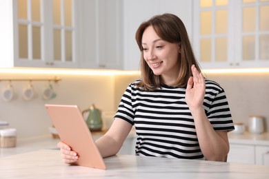 Photo of Woman having video chat via modern tablet at white marble table in kitchen