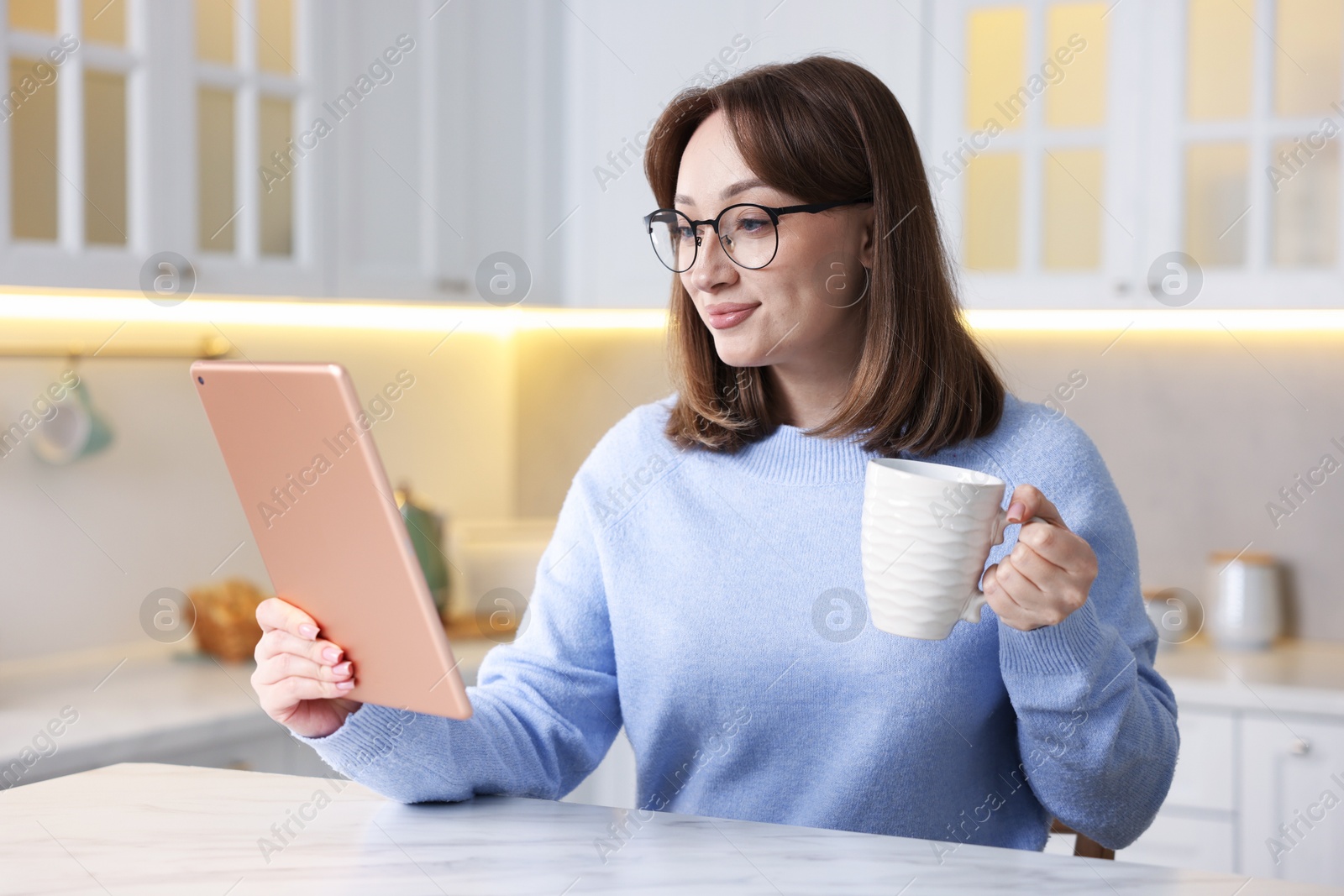 Photo of Woman with modern tablet and cup of drink at white marble table in kitchen