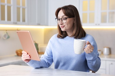 Photo of Woman with modern tablet and cup of drink at white marble table in kitchen