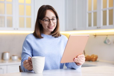 Photo of Woman with modern tablet and cup of drink at white marble table in kitchen