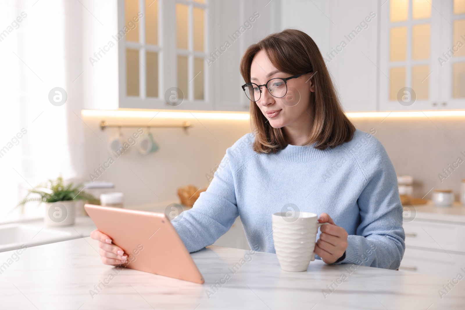 Photo of Woman with modern tablet and cup of drink at white marble table in kitchen