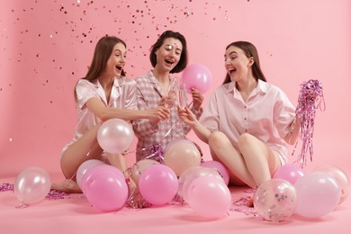 Photo of Pajama party. Happy friends with balloons and tinsel clinking glasses under falling confetti on pink background